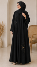 Woman wearing a black abaya with intricate designs in a room with a beige wall and wooden chair.